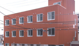 赤茶色の3階建て介護施設の建物外観。複数の窓とバスケットゴールが見える。